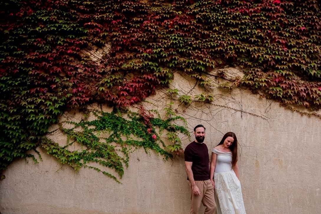 Lara y Javi de la mano junto a una fachada cubierta de hojas verdes y rojizas.