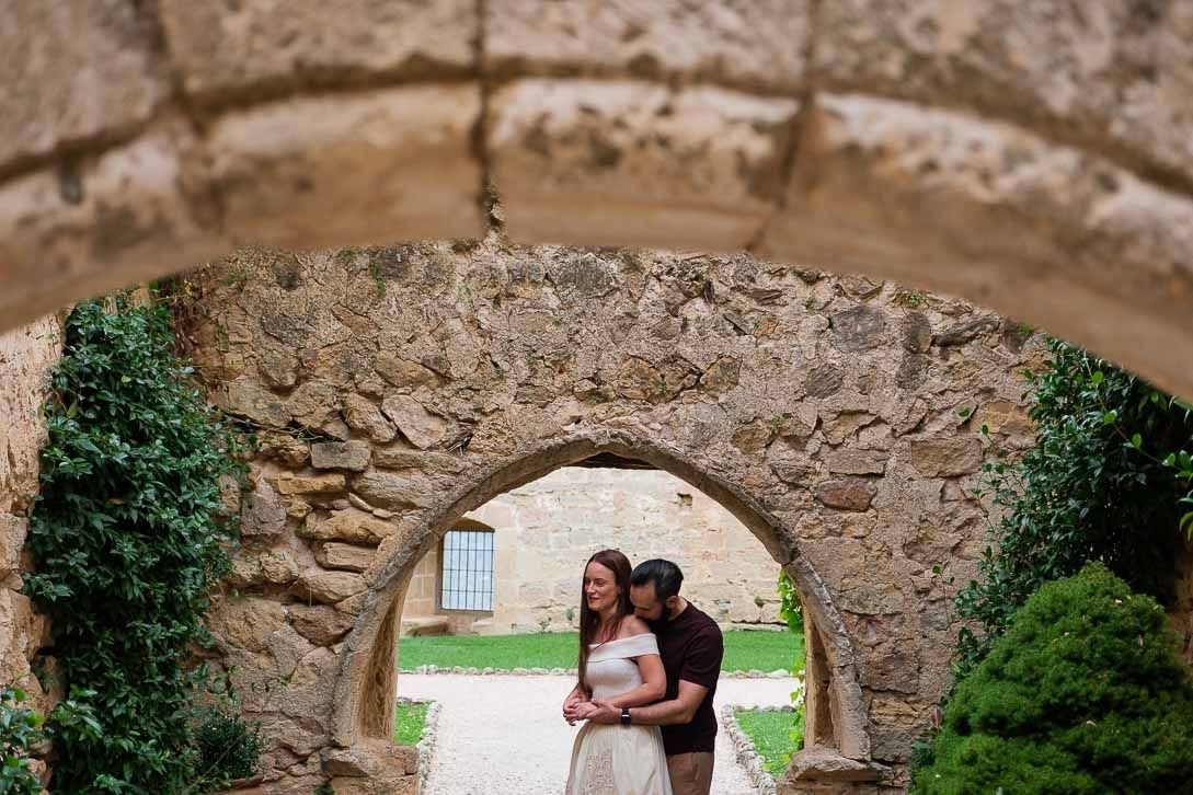 Javi abraza a Lara por detrás bajo un arco de piedra rodeado de vegetación.