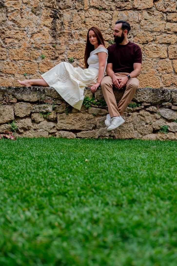 Retrato vertical de Lara y Javi sentados sobre un muro bajo, con césped en primer plano.