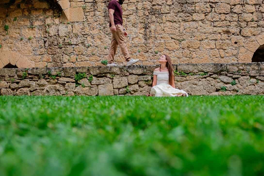 Lara sentada junto al césped mira a Javi, que camina sobre un muro bajo de piedra.
