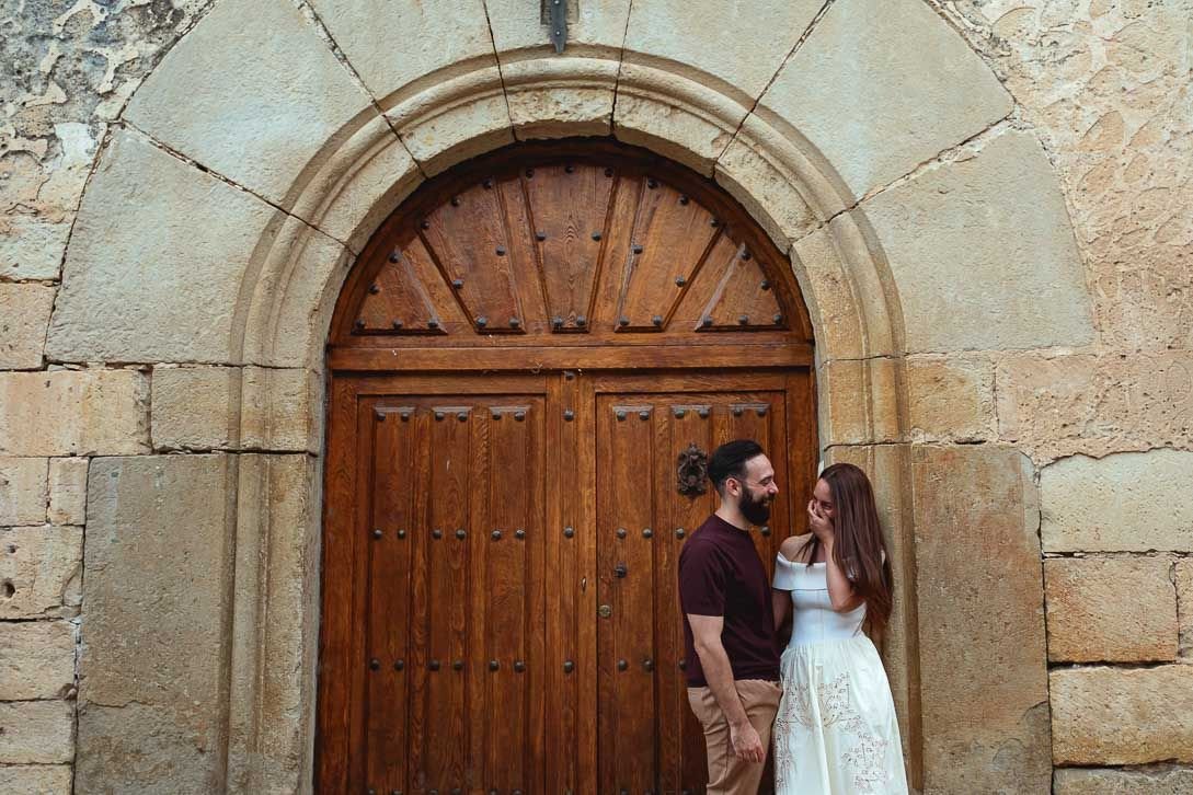 Lara ríe junto a Javi ante una gran puerta de madera con arco de piedra.