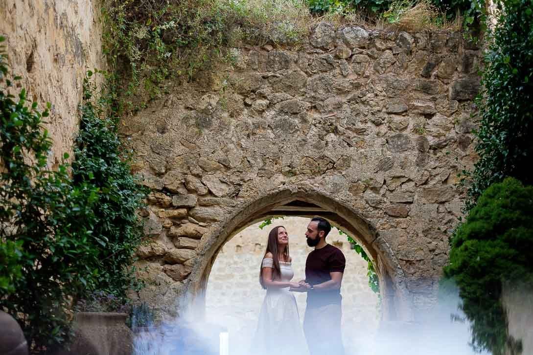 Lara y Javi de la mano bajo un arco de piedra en su preboda en Pedraza, con un reflejo claro en primer plano.